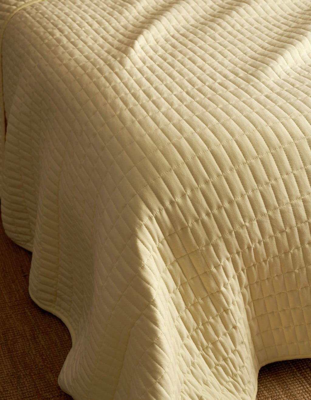A close-up of the Bamboo Jersey Quilt by Cozy Earth, featuring a cream color, quilted square pattern, and soft textured stitching draped over the edge of a bed.|Color:Meringue