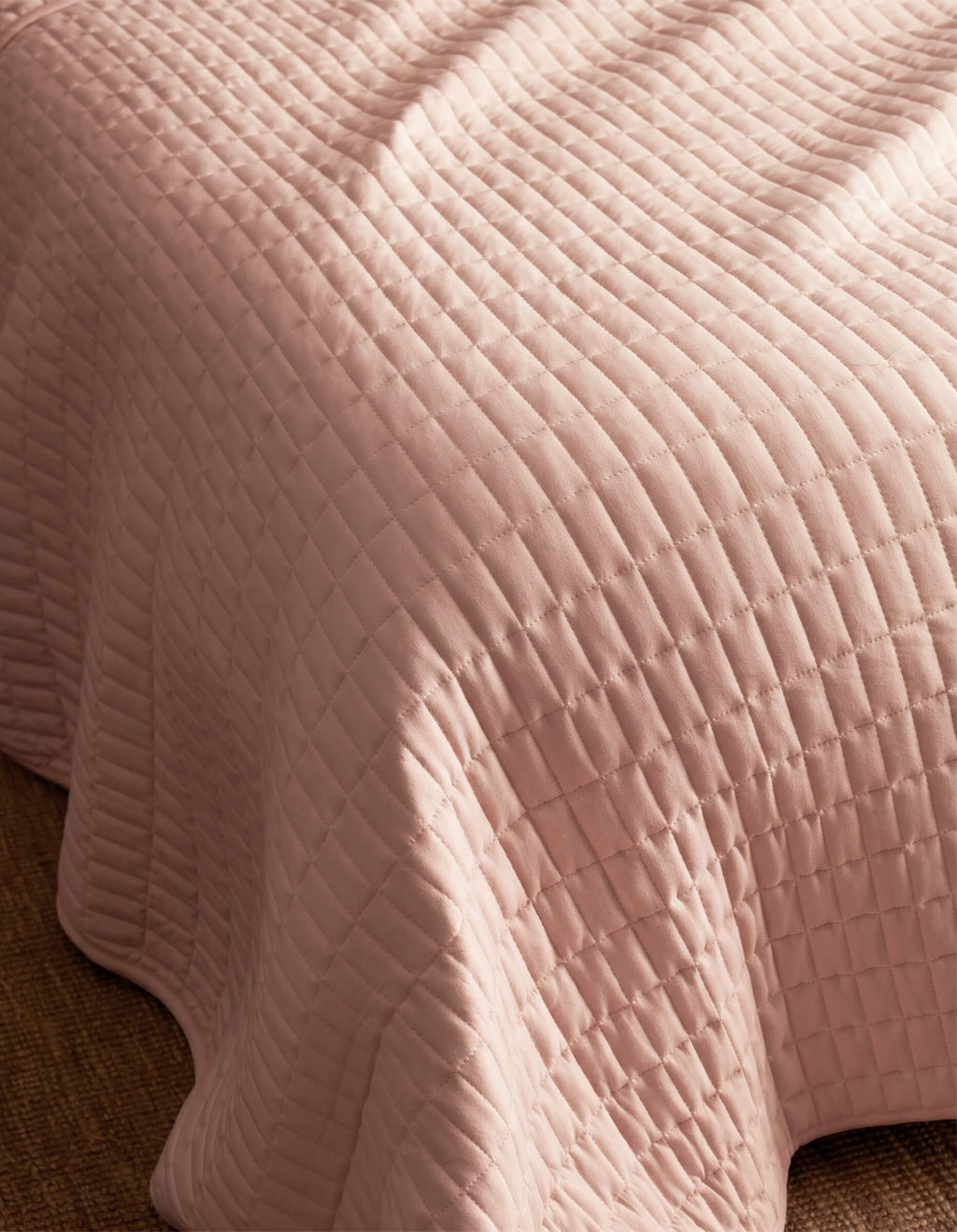 A close-up of the Cozy Earth Bamboo Jersey Quilt Bundle in pale pink with a quilted grid pattern, draped over a bed’s edge and resting on a textured brown carpet.|Color:Petal
