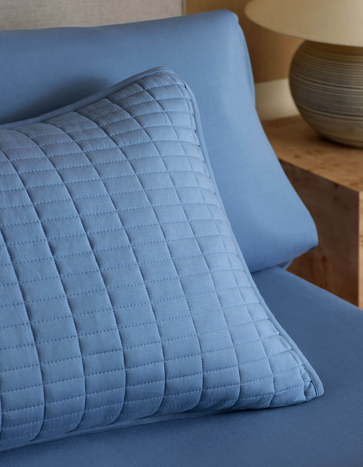 A Cozy Earth Bamboo Jersey Quilted Sham in blue rests on matching sheets, with a wooden nightstand and ceramic lamp visible in the background.|Color:Sailor