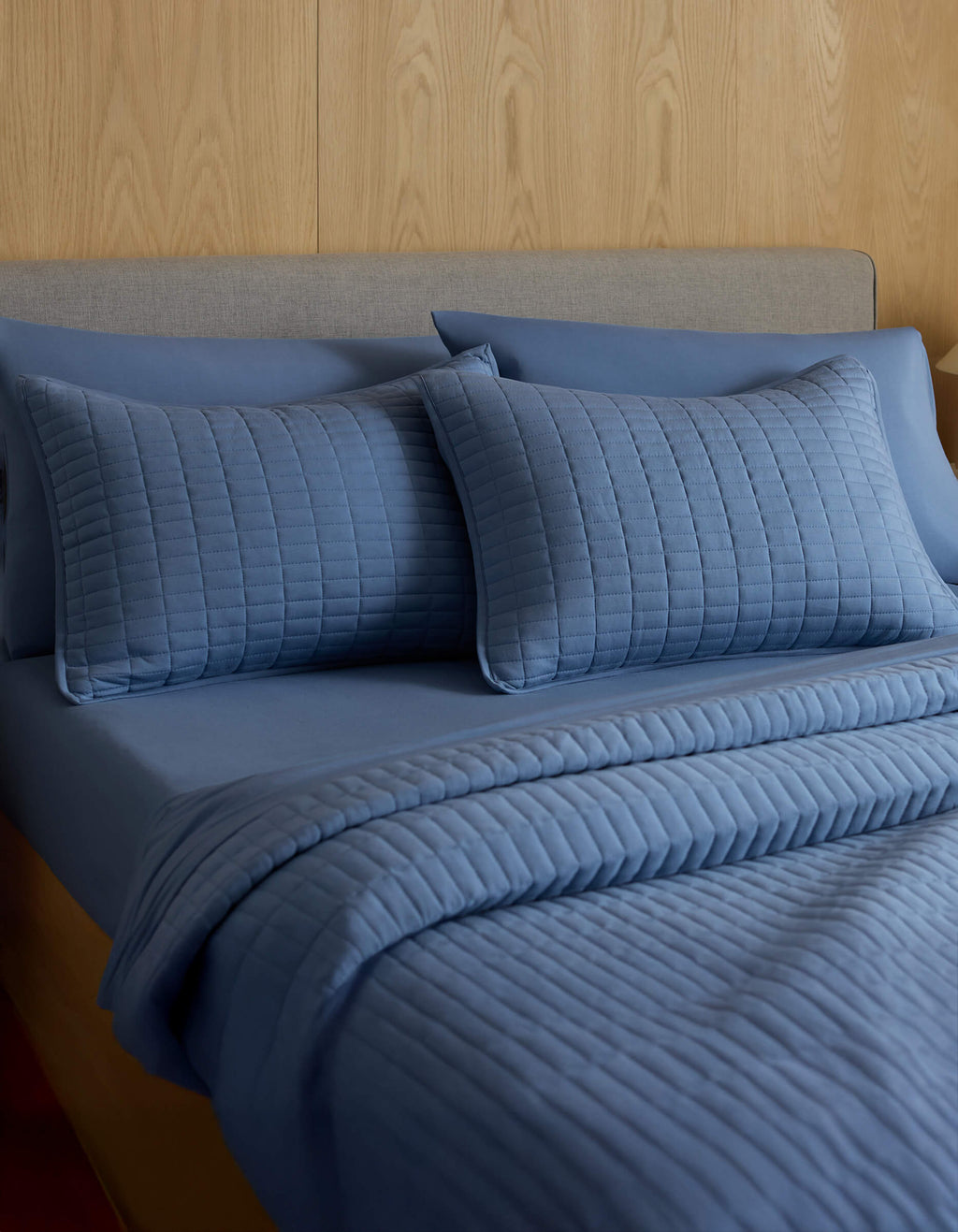 A neatly made bed with a grey headboard features Cozy Earth's Bamboo Jersey Quilted Shams in blue, a matching fitted sheet, and comforter, all set against a light wood-paneled wall.|Color:Sailor