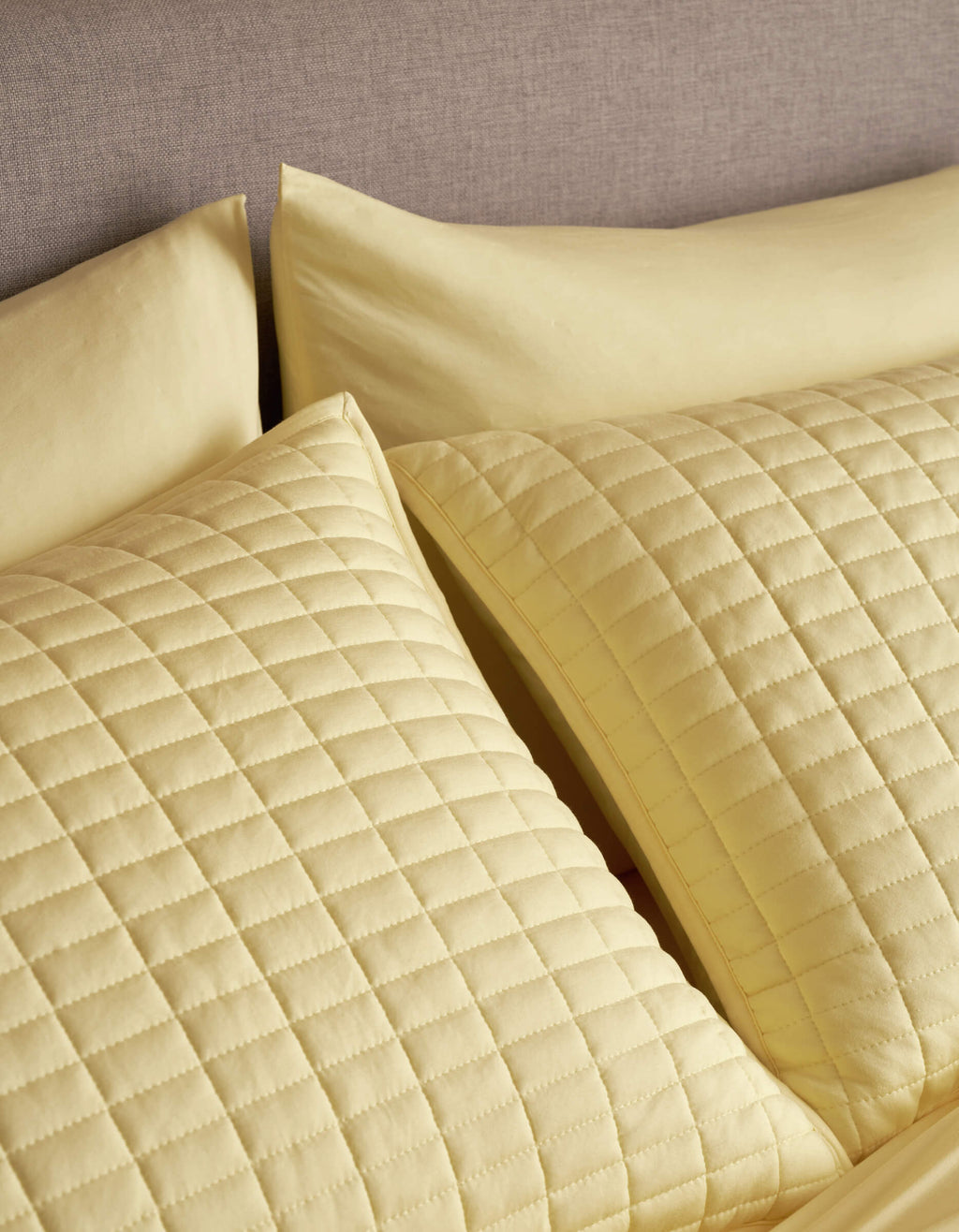 Two Cozy Earth Bamboo Jersey Quilted Shams in yellow, featuring a square pattern, rest against a grey upholstered headboard, paired with matching yellow bedsheets and pillowcases.|Color:Meringue