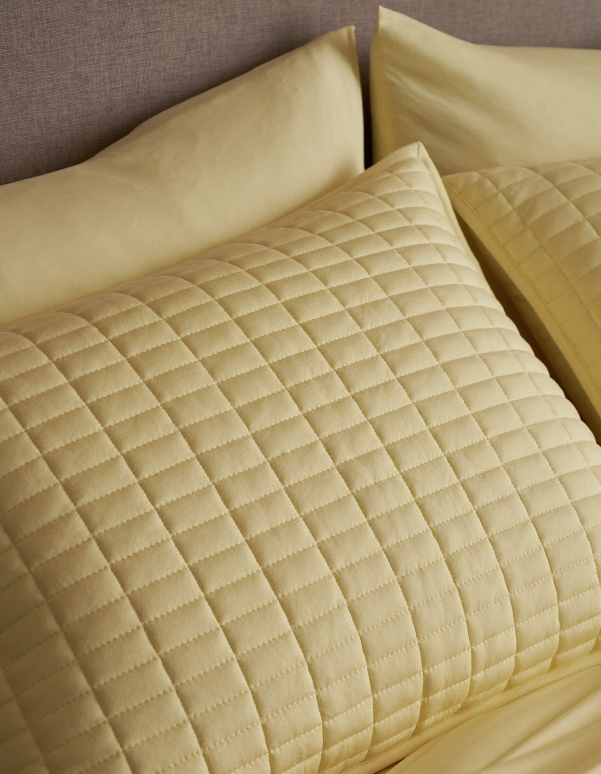 A close-up of a neatly made bed showcases Cozy Earth's Bamboo Jersey Quilted Shams in light yellow, featuring a square pattern and soft, textured appearance that matches the sheets.|Color:Meringue