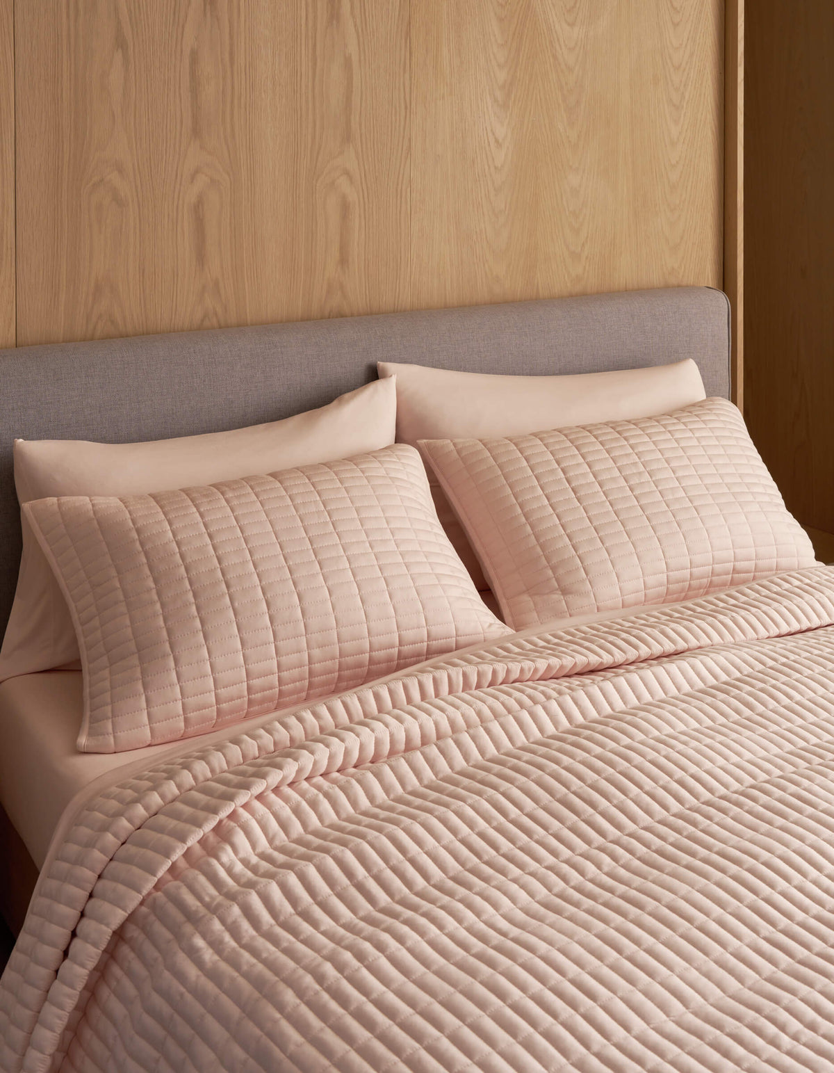 A neatly made bed features the Cozy Earth Bamboo Jersey Quilt in light pink with matching pillows, set against a light wood-paneled wall and a light grey upholstered headboard.|Color:Petal