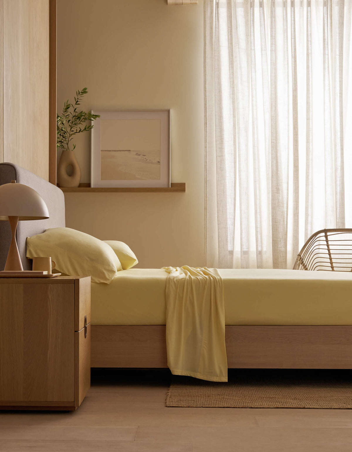 A minimal bedroom with a wooden bed frame, Cozy Earth's Bamboo Jersey Sheet Set in pale yellow, a wooden nightstand and lamp, a plant, framed art on a shelf, and sheer curtains letting in natural light.|Color:Meringue
