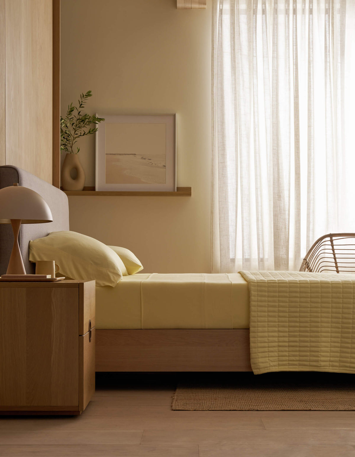 A minimal bedroom features a single bed with Cozy Earth’s Bamboo Jersey Flat Sheet in light yellow, a wooden nightstand holding a lamp and vase, a framed picture on a ledge, and sheer curtains letting in soft daylight.|Color:Meringue