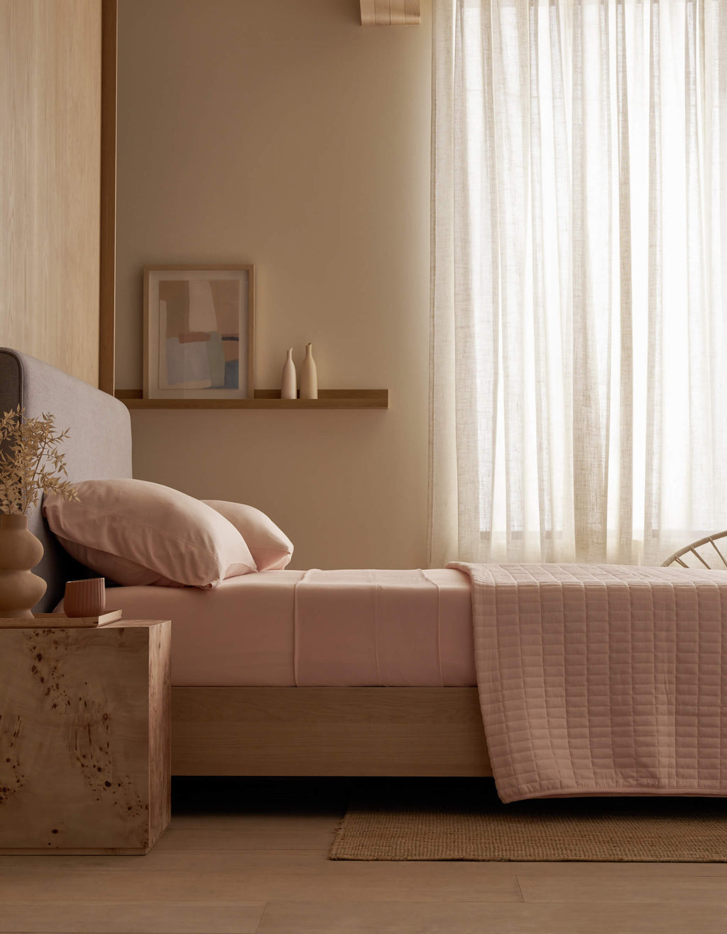 A modern bedroom featuring a bed with Cozy Earth Bamboo Jersey Fitted Sheet in pink, a wooden nightstand with books and a cup, a beige chair, potted plant, sheer curtains, and abstract blue wall art.|Color:Petal