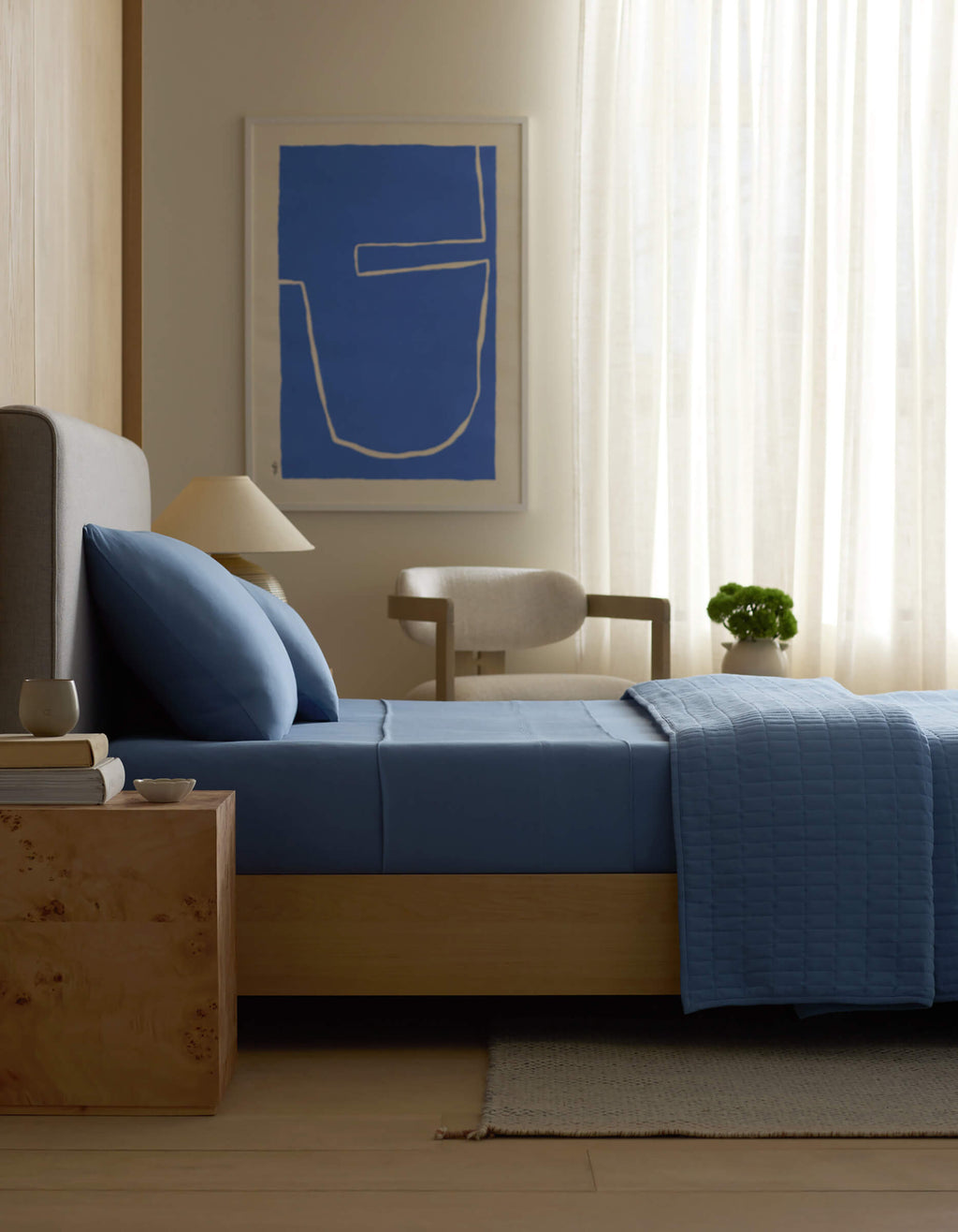 A modern bedroom featuring a bed with Cozy Earth Bamboo Jersey Fitted Sheet in blue, a wooden nightstand with books and a cup, a beige chair, potted plant, sheer curtains, and abstract blue wall art.|Color:Sailor