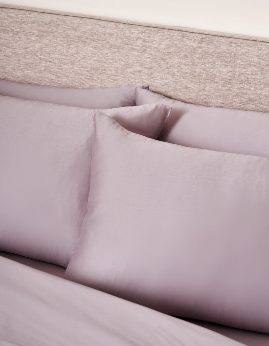 Two Cozy Earth Bamboo Pillowcases in lavender rest against a matching headboard, with a coordinating lavender sheet partially visible beneath them. The bedding and pillowcases feature a smooth, soft texture.|Color:Lavender