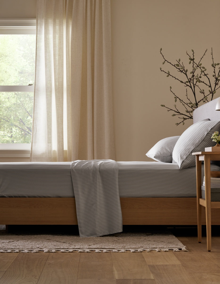 A bed neatly made with the Cozy Earth Bamboo Flat Sheet in gray stripes sits beside a wooden nightstand, set in a softly lit room with light curtains, a window, and a decorative plant branch in the background.|Color:Classic Stripe in Dew