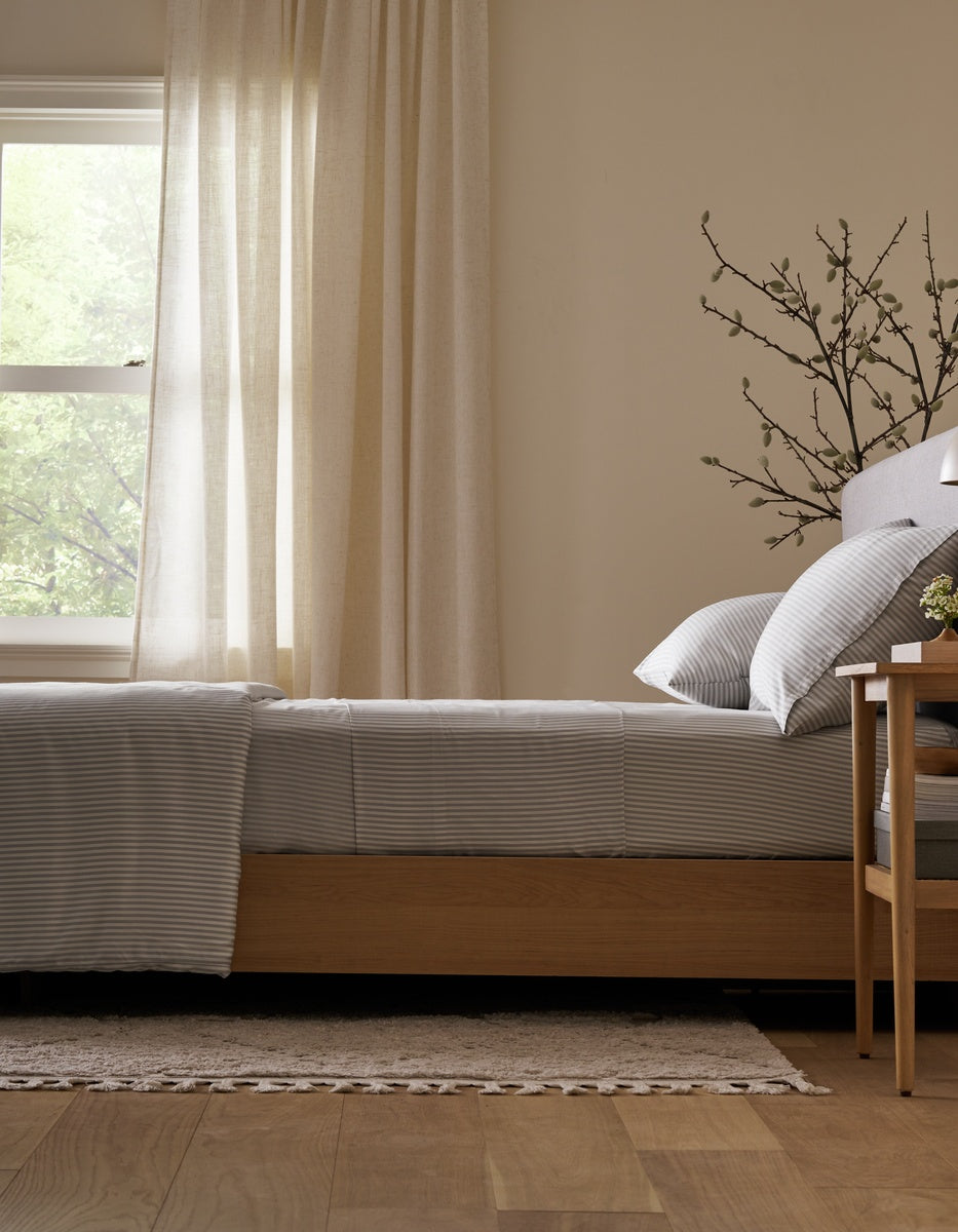 A neatly made bed featuring the Cozy Earth Bamboo Fitted Sheet sits beside a wooden nightstand, a branch in a vase, and a window with sheer curtains letting in natural light. Light wood floors and a rug complete the cozy scene.|Color:Classic Stripe in Dew