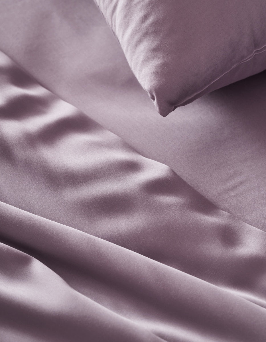 A close-up of a smooth, shiny Bamboo Flat Sheet and matching pillowcase by Cozy Earth in lavender, showcasing soft folds and a luxurious texture.|Color:Lavender