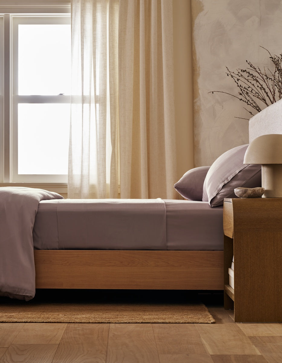 A neatly made bed with Cozy Earth’s Bamboo Fitted Sheet sits beside a wooden nightstand adorned with a lamp and dried branches, while light curtains allow sunlight to filter in through the window.|Color:Lavender
