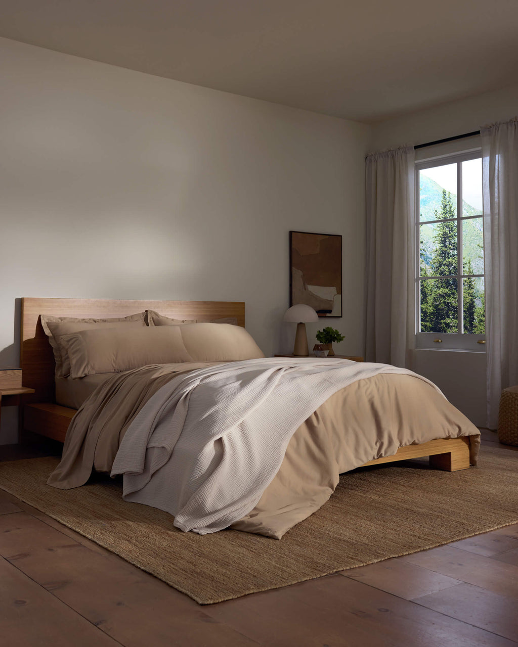 A cozy bedroom features a wooden bed with beige bedding and a white Cotton Gauze Blanket by Cozy Earth, a bedside table with a lamp, sheer curtains letting in natural light, and a woven rug on the wooden floor.|Color:Coconut