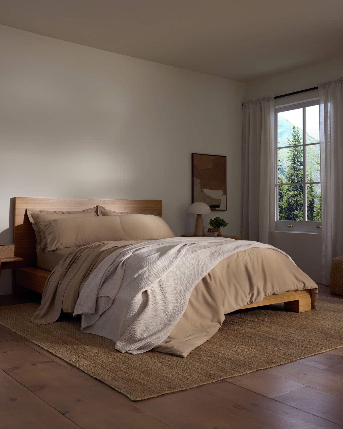 A cozy bedroom features a wooden bed with beige bedding and a white Cotton Gauze Blanket by Cozy Earth, a bedside table with a lamp, sheer curtains letting in natural light, and a woven rug on the wooden floor.|Color:Coconut