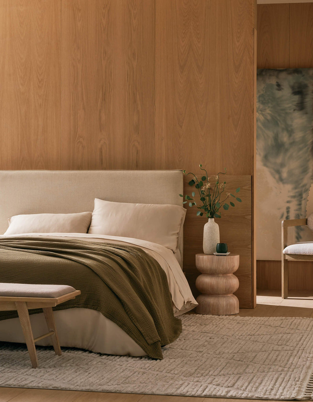 A modern, minimalist bedroom with a beige bed, Cozy Earth’s Cotton Gauze Blanket in olive green, light wooden walls, a textured rug, sculptural nightstand with a small vase of greenery, cushioned chair, and abstract wall art.|Color:Sable