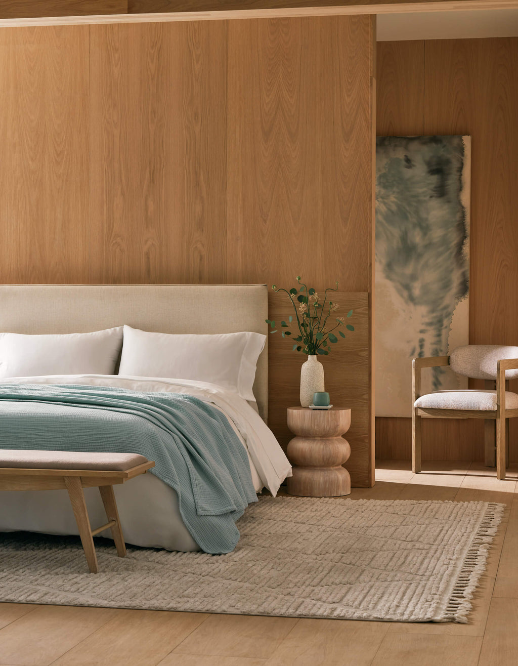 A modern bedroom with light wood-paneled walls, a cream bed topped with Cozy Earth's Cotton Gauze Blanket, white and light blue bedding, a layered side table with greenery, a beige bench, and a soft woven rug.|Color:Tide