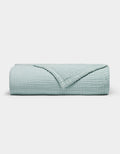 The Cozy Earth Cotton Gauze Blanket is neatly folded in light blue with a textured, grid-like pattern on a plain white background. One corner is turned down to reveal the inner side.|Color:Tide