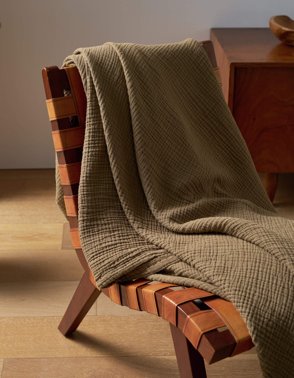 A Cozy Earth Cotton Gauze Blanket in olive green is draped over a wooden chair with a woven leather seat, set on a light wood floor beside another wooden furniture piece.|Color:Sable