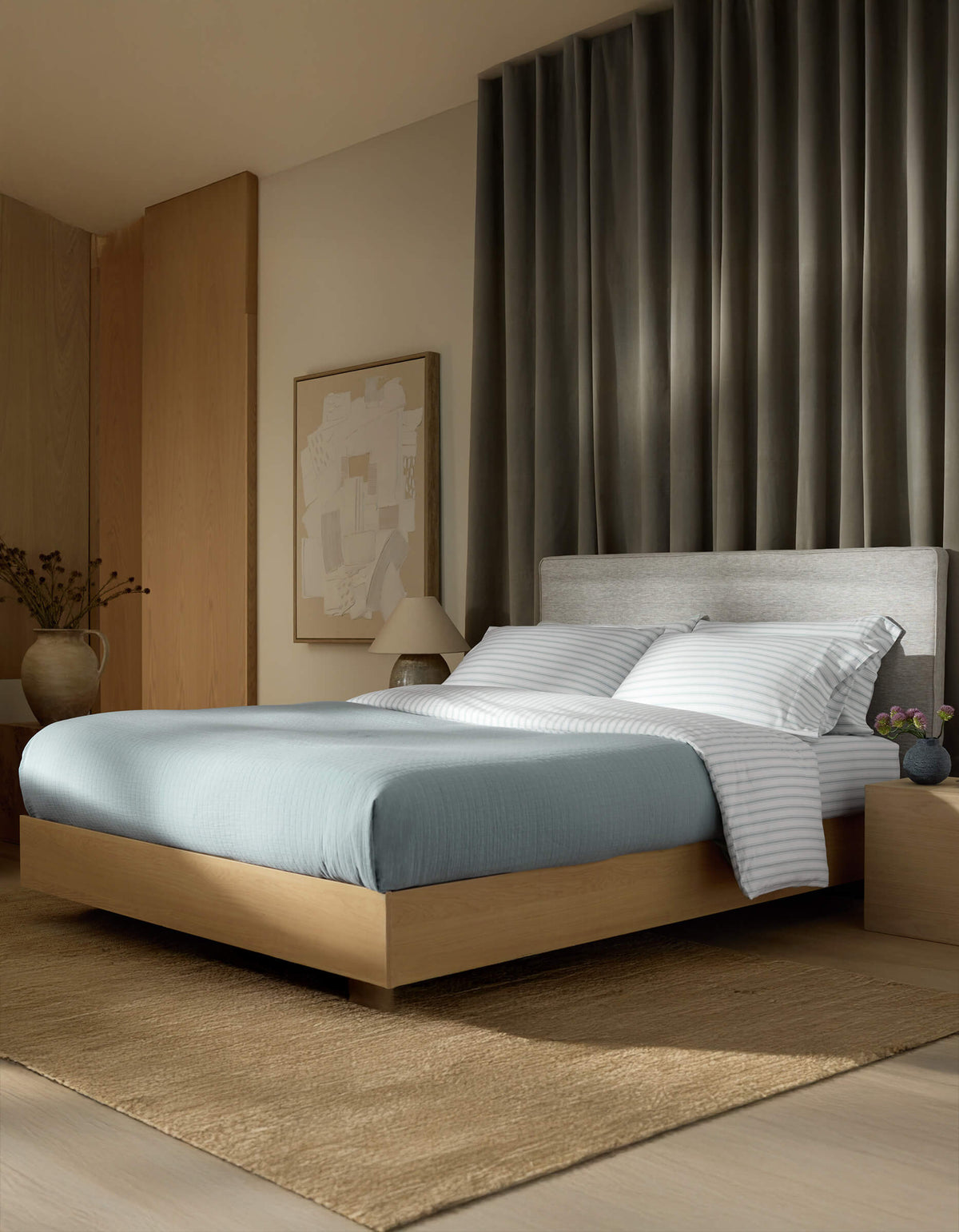A modern bedroom features Cozy Earth’s Waterfront Grid Matelasse Blanket in blue, a light wood platform bed, striped white pillows, gray upholstered headboard, neutral rug, nightstand with vase, large abstract wall art, and dark curtains behind the bed.|Color:River