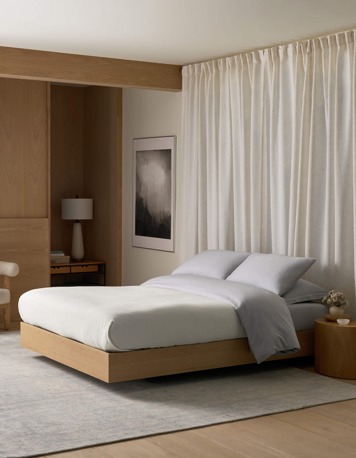 A modern, minimalist bedroom featuring a light wood platform bed, the Cozy Earth Waterfront Grid Matelasse Blanket in gray, a round wooden nightstand, lamp, abstract wall art, light curtains, and soft natural lighting.|Color:Sea Salt