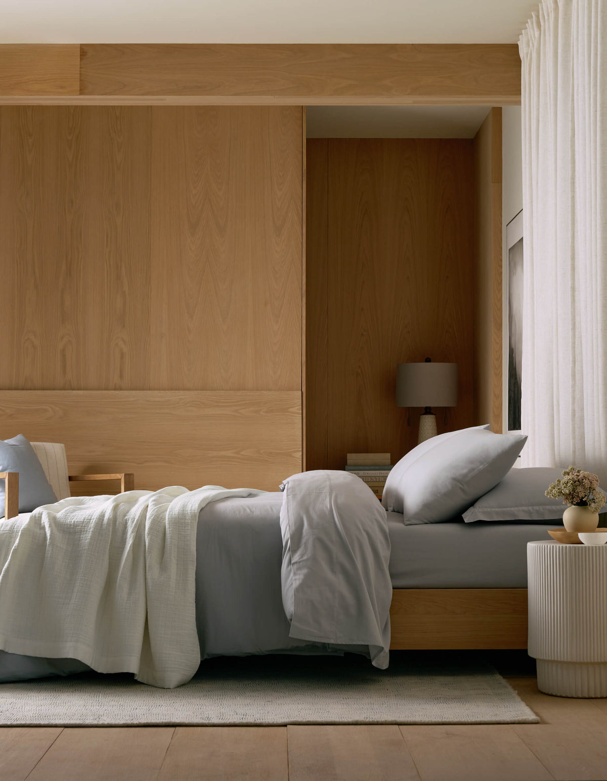 A modern, minimalist bedroom with a neatly made bed featuring Cozy Earth’s Waterfront Grid Matelasse Blanket in light gray, a wooden headboard, table lamp, round white side table with flowers, light wood paneling, and cream curtains.|Color:Sea Salt