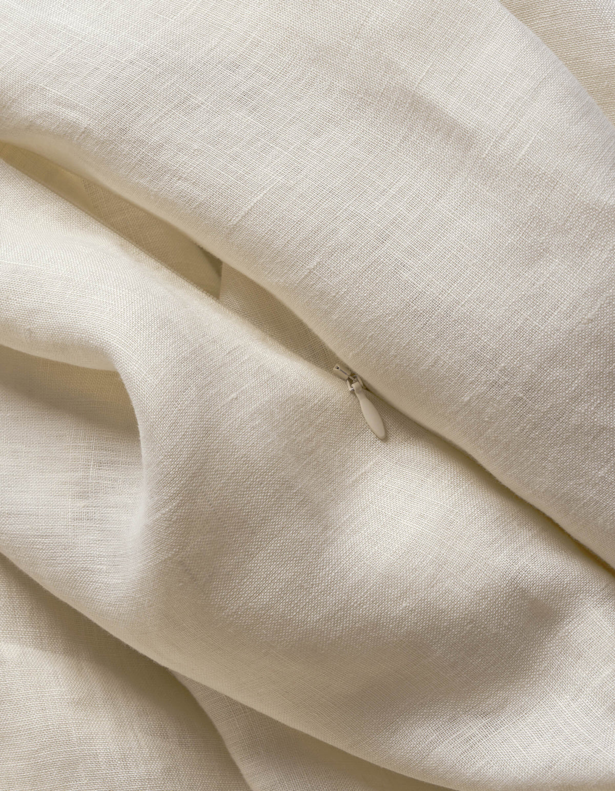 A cream-colored Cozy Earth Linen Duvet Cover with a textured weave is softly draped and folded, revealing a small, partially visible metal zipper pull among the folds.|Color:Flax