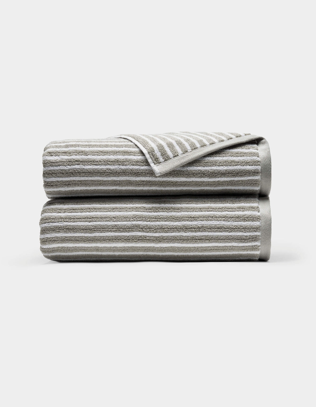 Two neatly folded Ribbed Terry Bath Towels by Cozy Earth, light gray with horizontal white stripes, stacked atop each other against a plain white background. A corner of the top towel is turned over, revealing the underside.|Color:Dew Stripe