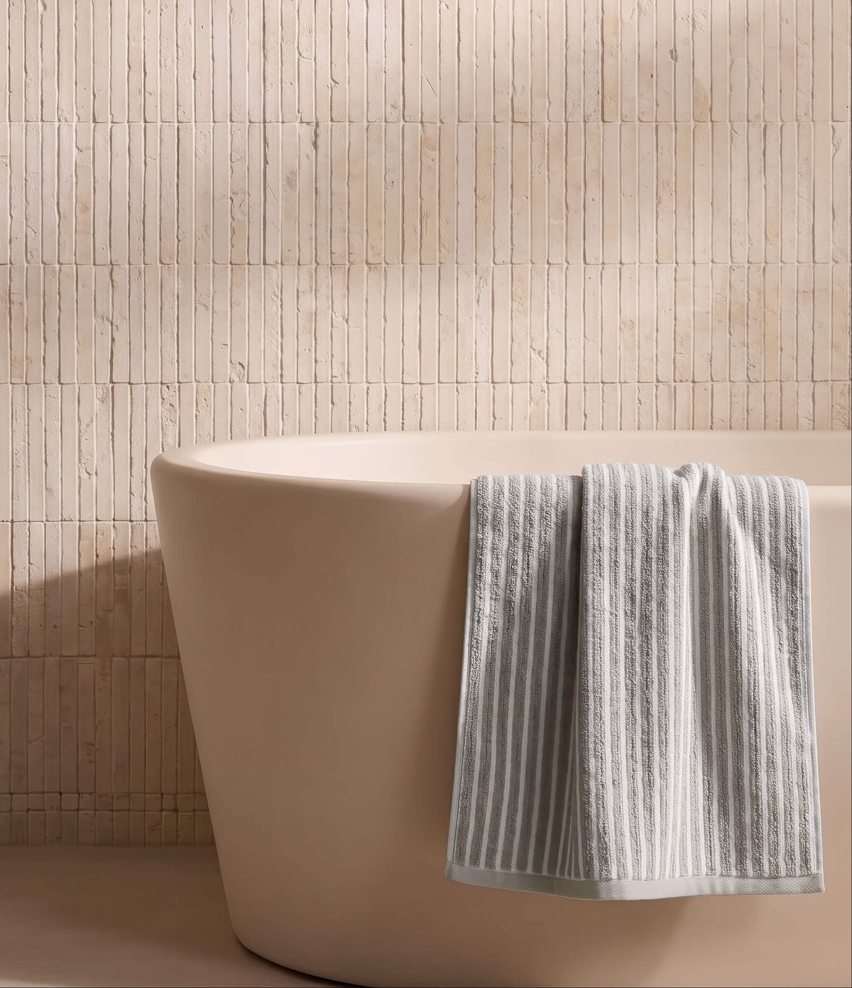 A beige freestanding bathtub with a Cozy Earth Ribbed Terry Bath Towel draped over the side, set against a light tan tiled wall.|Color:Dew Stripe