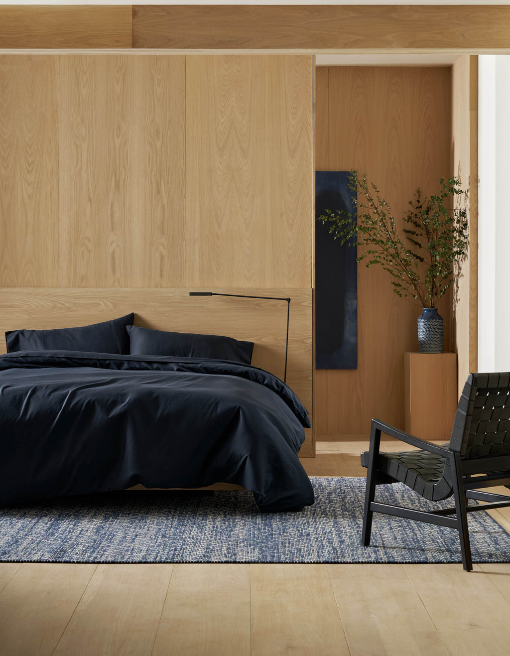 A modern bedroom with a wooden accent wall features the Soft-Wash Cotton Bedding Deluxe Bundle by Cozy Earth on the bed, a black floor lamp, a black woven chair, a blue rug, and a tall vase with branches on a wooden pedestal.|Color:Eclipse