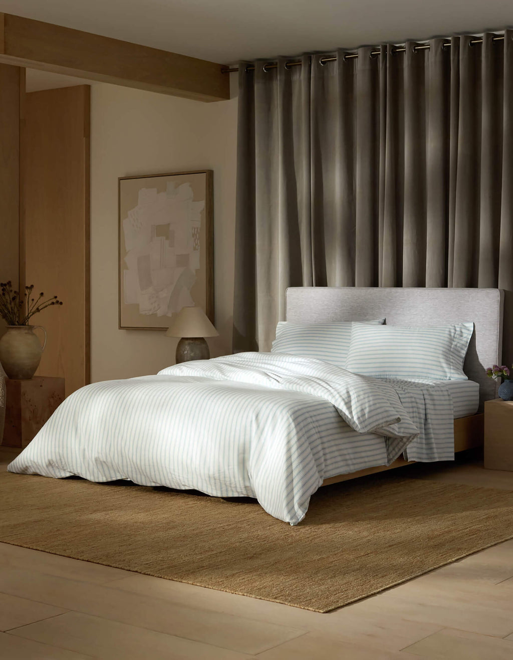 A modern bedroom with a neatly made bed featuring Cozy Earth's Soft-Wash Cotton Duvet Cover, a light headboard, brown textured rug, gray floor-length curtains, bedside table with a lamp and vase, and abstract wall art.|Color:River and Sea Salt Stripe