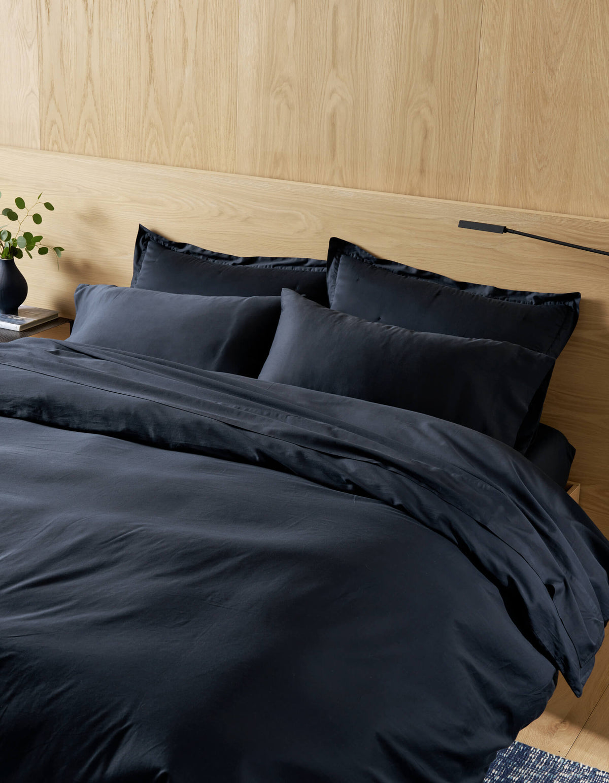A neatly made bed features Cozy Earth Soft-Wash Cotton Pillowcases in dark navy, along with matching bedding and a comforter, against a light wooden headboard. A vase with green stems rests on the bedside table.|Color:Eclipse