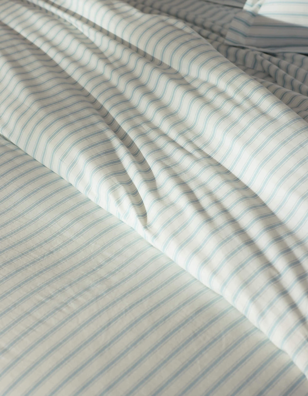 Close-up of a bed featuring the Cozy Earth Soft-Wash Cotton Duvet Cover in white and light blue stripes, gently wrinkled and illuminated by natural sunlight.|Color:River and Sea Salt Stripe