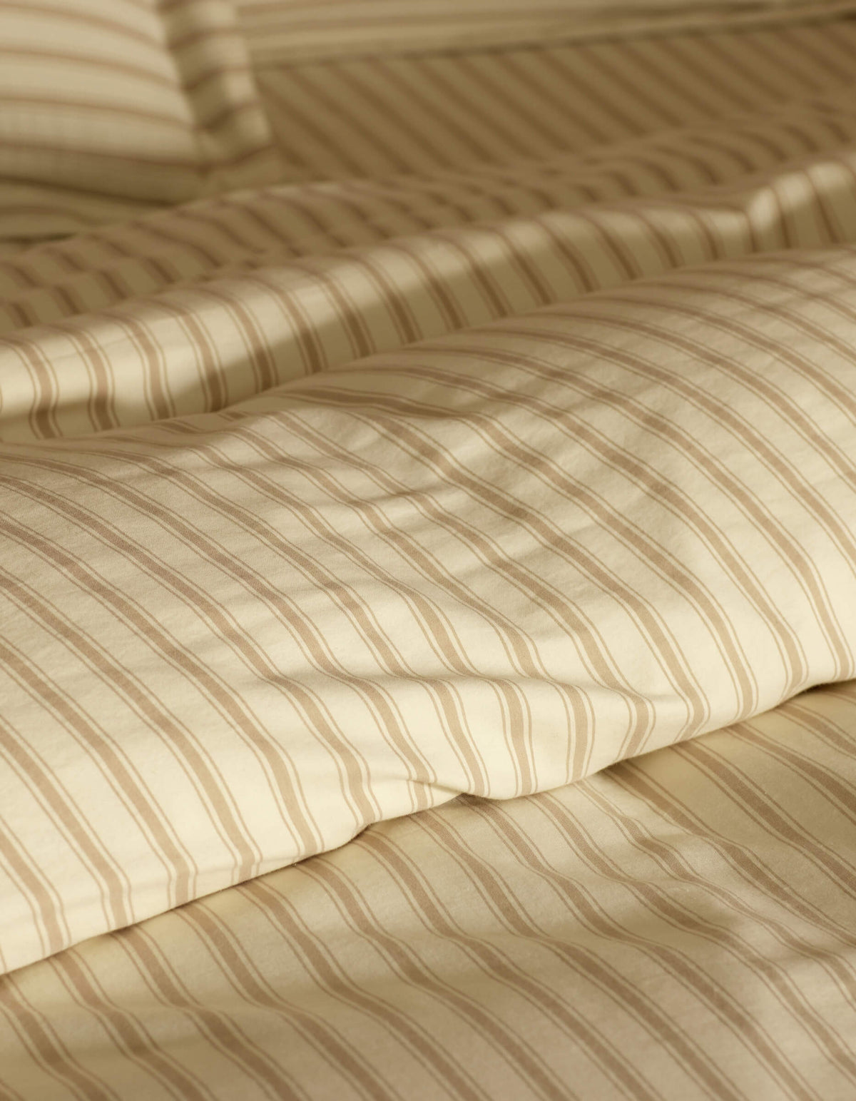 Close-up of a neatly made bed featuring Cozy Earth's Soft-Wash Cotton Duvet Cover in cream and beige stripes, with a matching pillow, softly lit to accentuate the smooth fabric texture.|Color:Coconut and Sandalwood Stripe