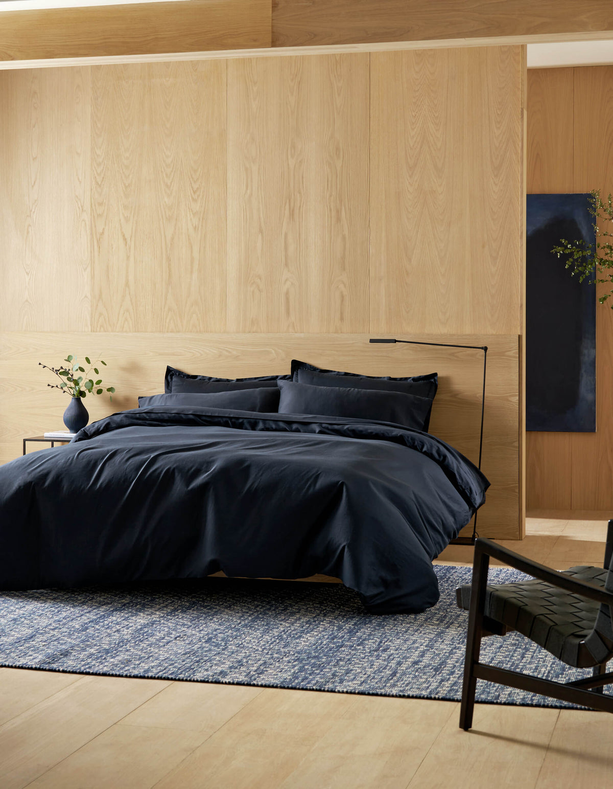 A modern bedroom features the Cozy Earth Soft-Wash Cotton Duvet Cover in black, with pillows, light wood walls and floors, a blue patterned rug, a black chair, and a small bedside table displaying a plant in a vase.|Color:Eclipse
