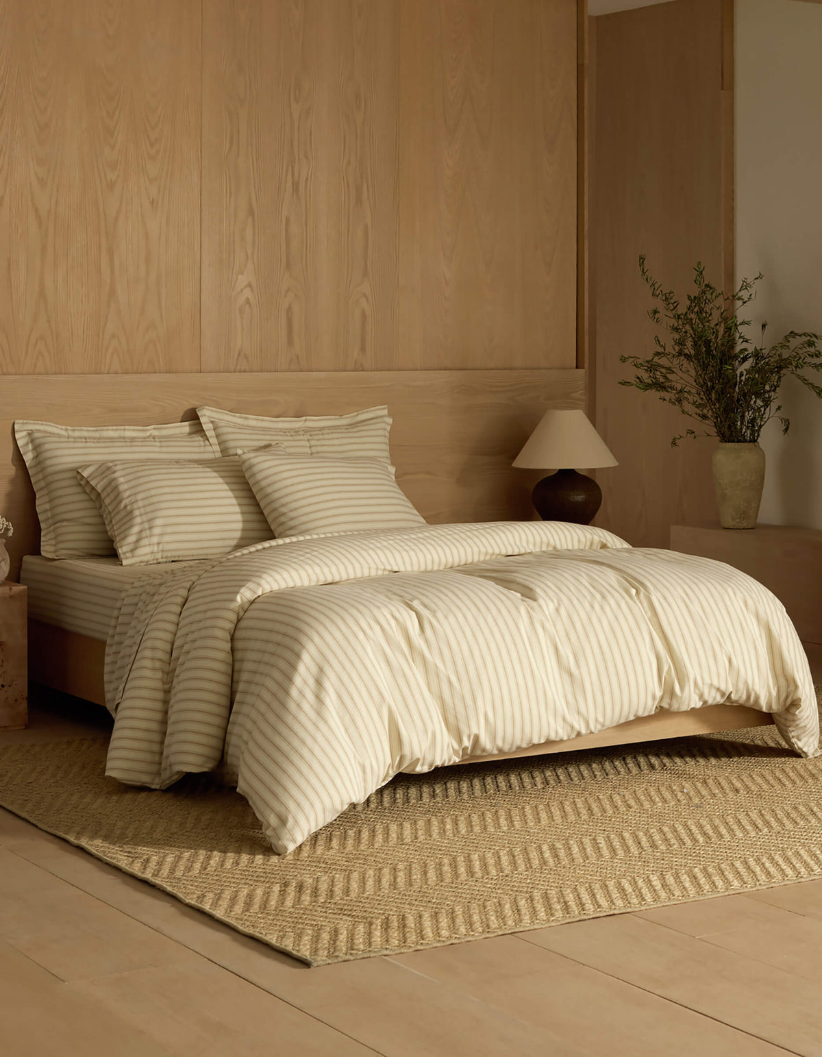 A minimalist bedroom features a neatly made bed dressed in the Cozy Earth Soft-Wash Cotton Duvet Cover, set atop a textured rug with light wood paneling, a nightstand, lamp, and a vase with greenery.|Color:Coconut and Sandalwood Stripe