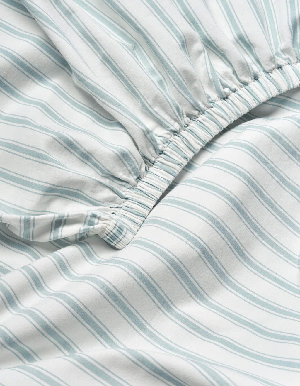 Close-up of the Soft-Wash Cotton Fitted Sheet by Cozy Earth, showing light blue and white vertical stripes and an elastic gathered edge for a snug fit.|Color:River and Sea Salt Stripe