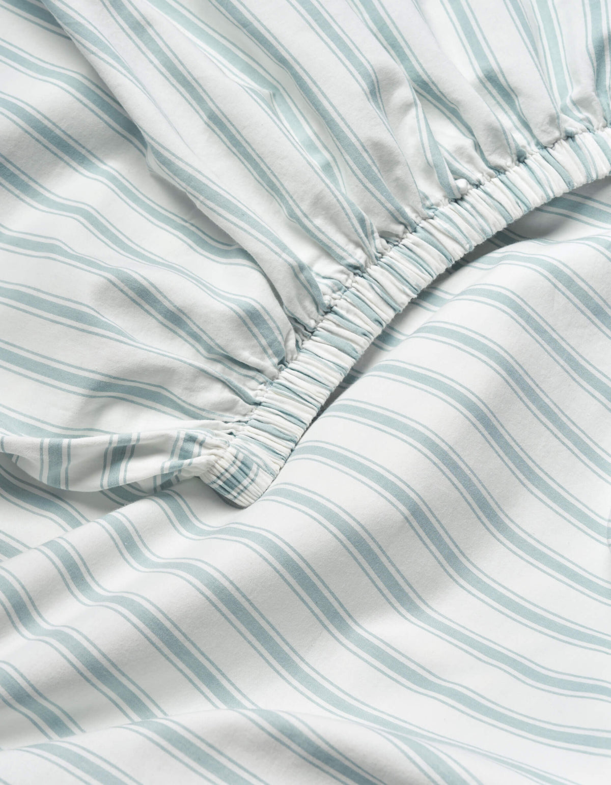 Close-up of the Soft-Wash Cotton Fitted Sheet by Cozy Earth, showing light blue and white vertical stripes and an elastic gathered edge for a snug fit.|Color:River and Sea Salt Stripe