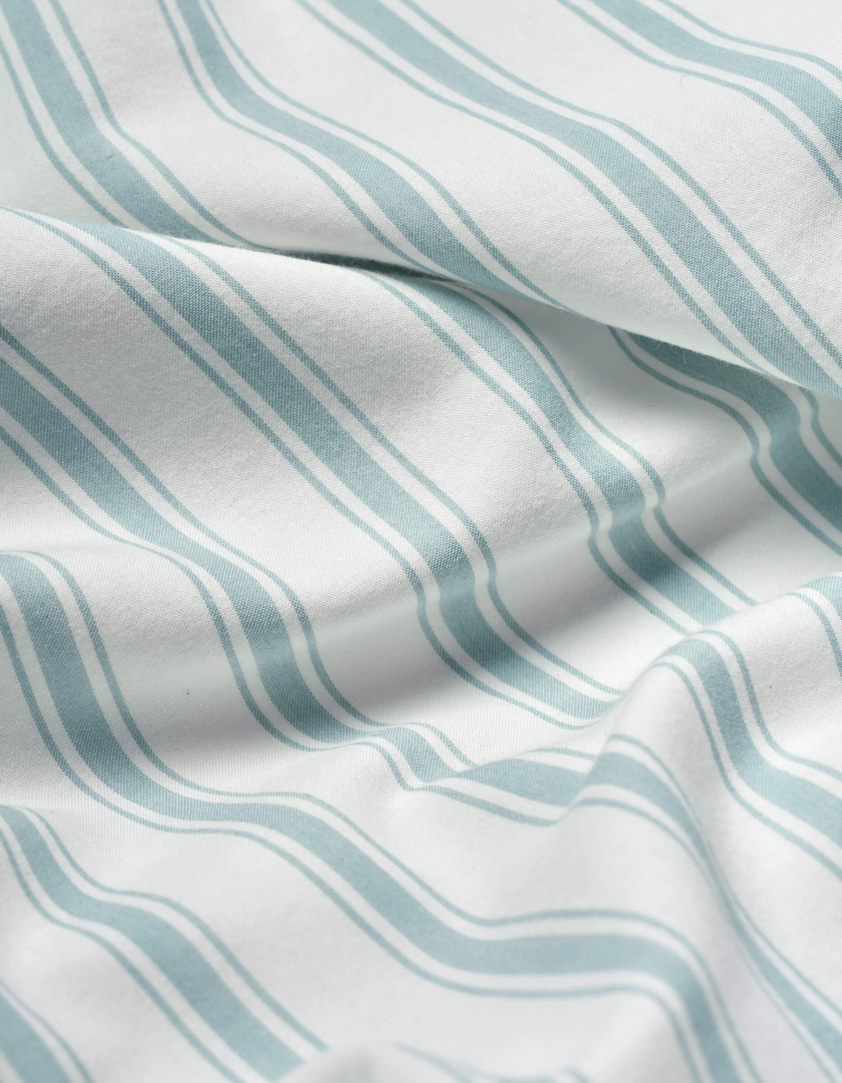 Close-up of Cozy Earth’s Soft-Wash Cotton Sheet Set in white, featuring light blue wavy parallel stripes. The soft fabric is slightly wrinkled, forming gentle folds and subtle shadows.|Color:River and Sea Salt Stripe