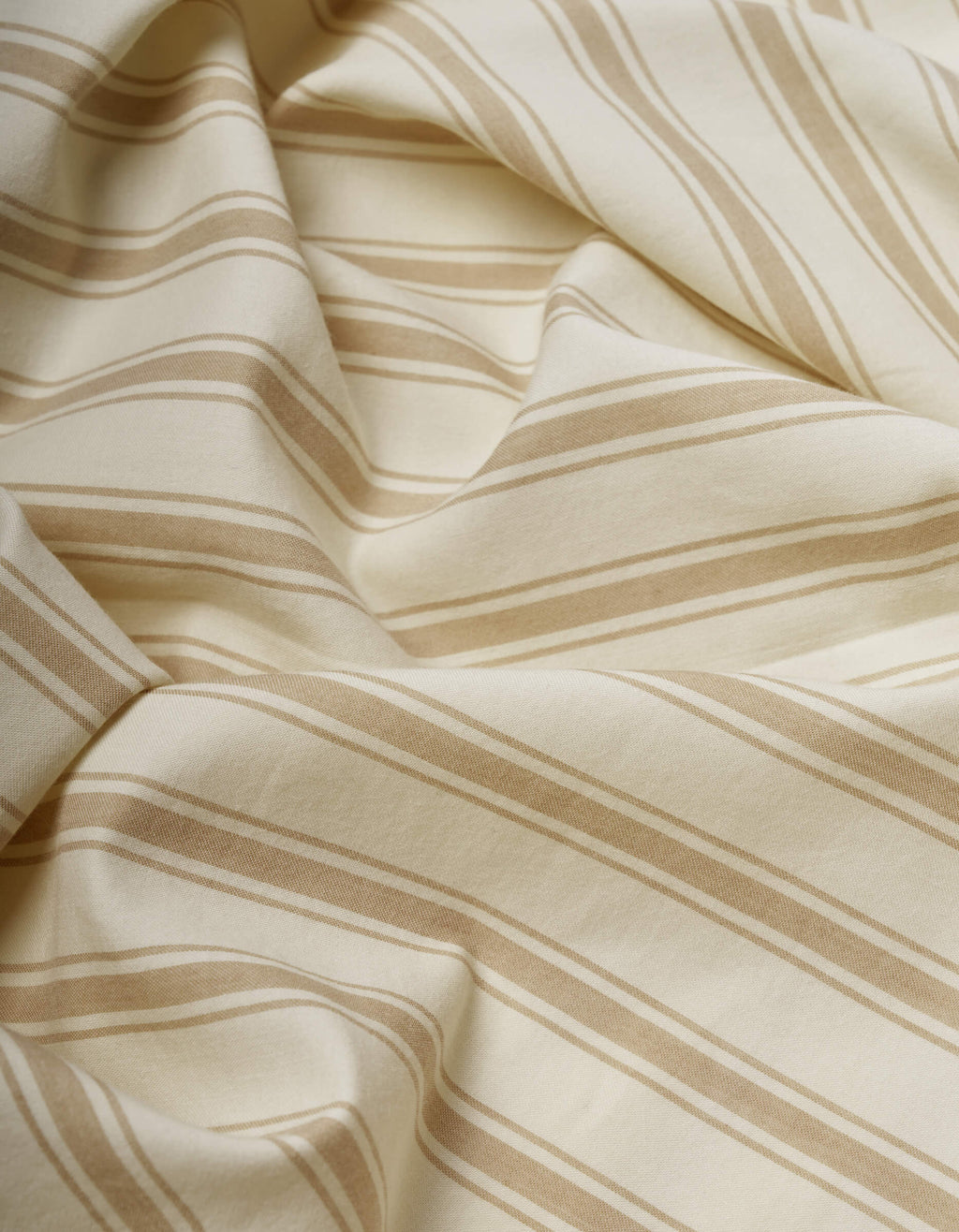 A close-up of Cozy Earth's Soft-Wash Cotton Sheet Set in cream with beige diagonal stripes, softly draped and folded to reveal gentle curves and subtle shadows.|Color:Coconut and Sandalwood Stripe