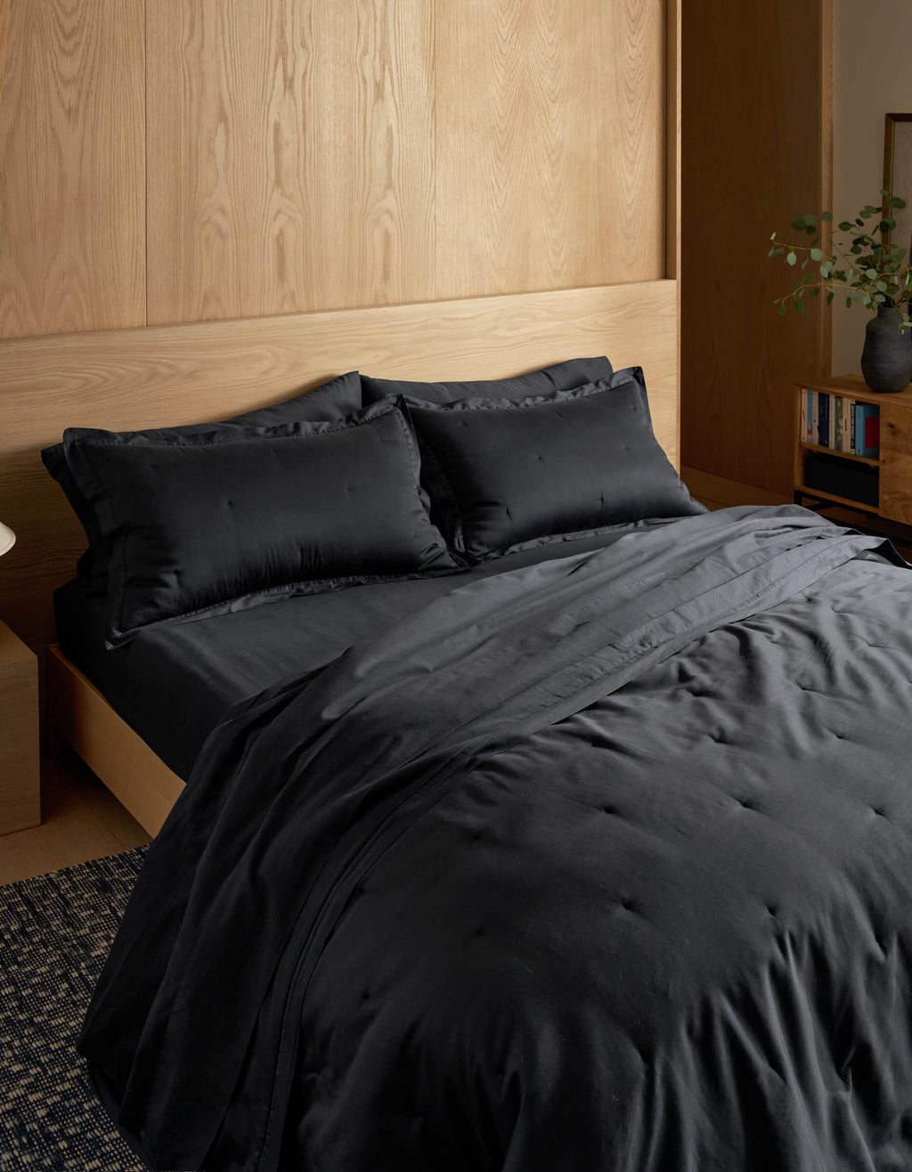 A modern, minimalist room features a neatly made bed with Cozy Earth’s Soft-Wash Cotton Quilt in black, set against a light wood headboard and wall. In the corner, a bookshelf holds books and a vase with greenery.|Color:Eclipse