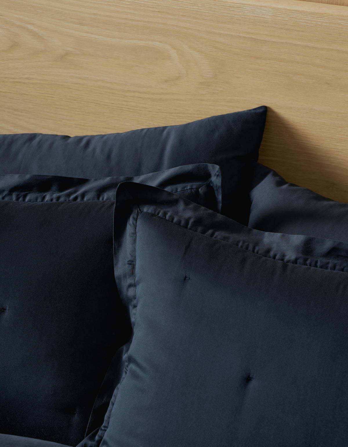 Two Soft-Wash Cotton Quilted Shams from Cozy Earth rest against a light wood headboard, with a black duvet partially visible in the foreground.|Color:Eclipse