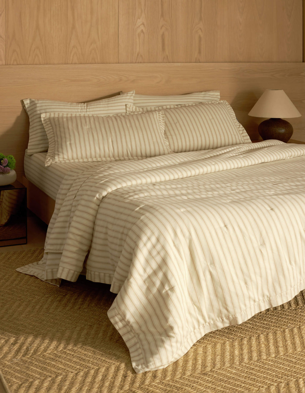 A bed featuring Cozy Earth's Soft-Wash Cotton Quilted Shams sits against a wooden paneled wall, complemented by matching pillows and a comforter. A bedside table with a lamp and vase, plus a woven rug on the floor, complete the cozy scene.|Color:Coconut and Sandalwood Stripe