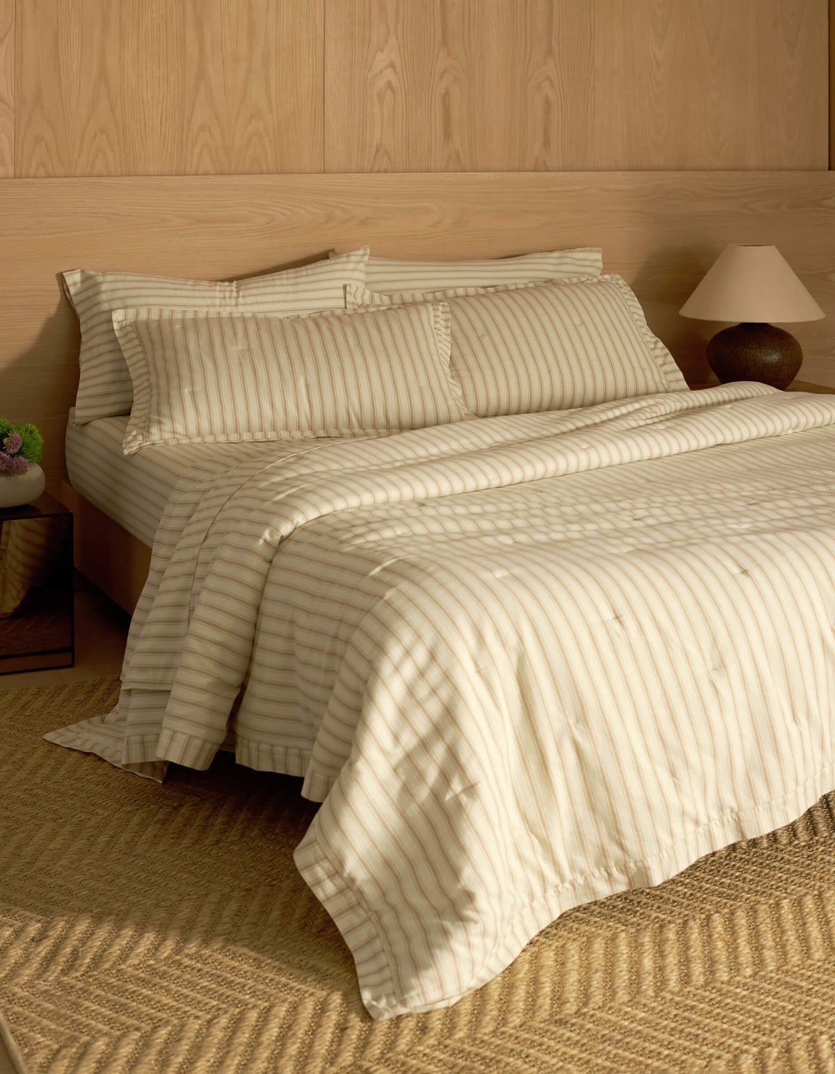 A bed featuring Cozy Earth's Soft-Wash Cotton Quilted Shams sits against a wooden paneled wall, complemented by matching pillows and a comforter. A bedside table with a lamp and vase, plus a woven rug on the floor, complete the cozy scene.|Color:Coconut and Sandalwood Stripe