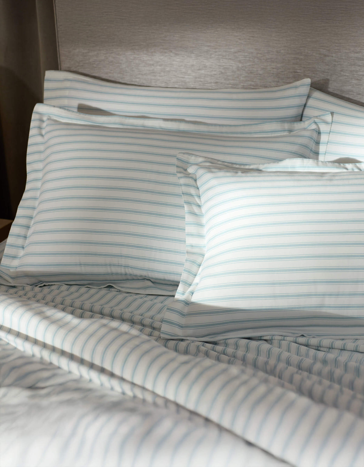 A bed featuring Cozy Earth Soft-Wash Cotton Shams in white and light blue stripes is set against a gray upholstered headboard, softly illuminated by sunlight.|Color:River and Sea Salt Stripe