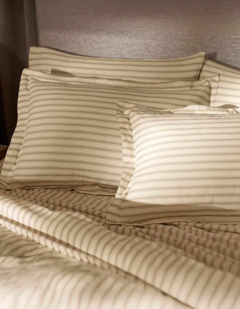 A bed featuring Cozy Earth Soft-Wash Cotton Shams in tan and beige stripes is set against a gray upholstered headboard, softly illuminated by sunlight.|Color:Coconut and Sandalwood Stripe