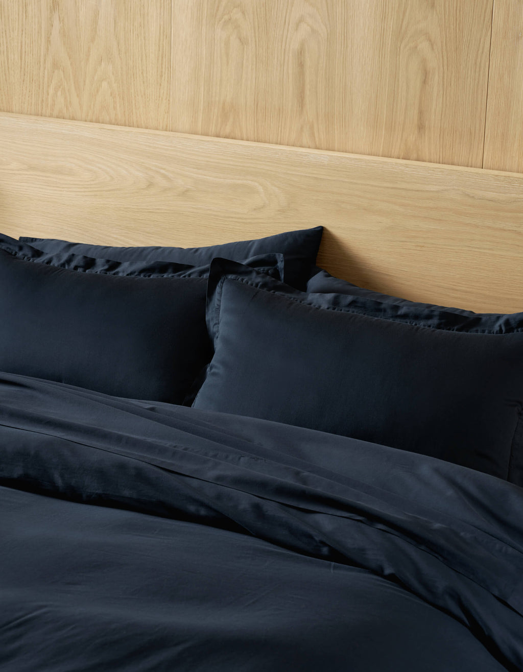 A neatly made bed with Cozy Earth Soft-Wash Cotton Shams in dark navy, paired with matching sheets, stands against a light wooden headboard and a wood-paneled wall.|Color:Eclipse