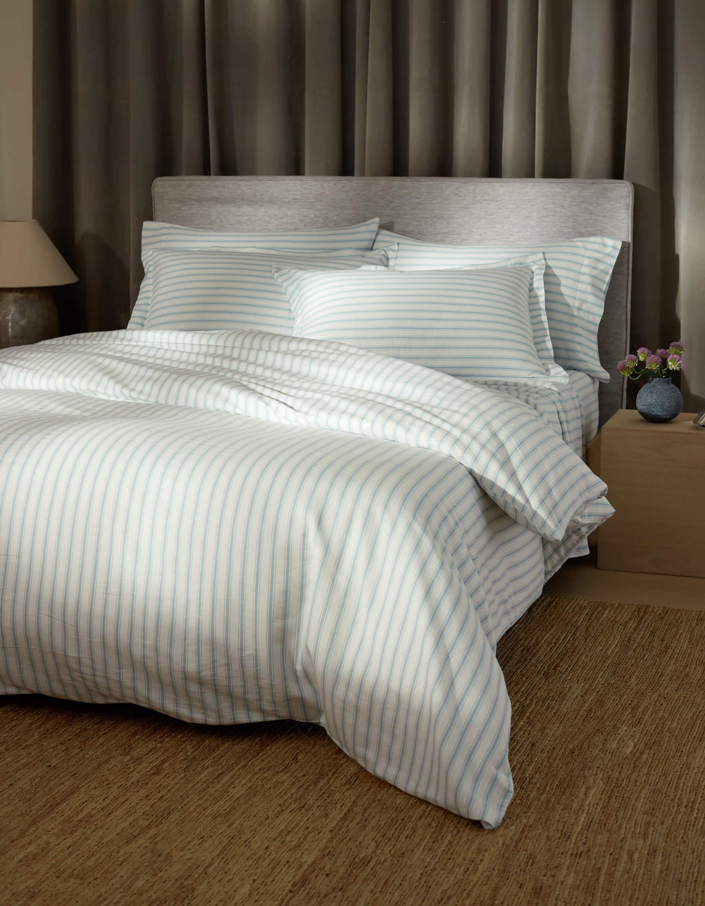 A bed features Cozy Earth Soft-Wash Cotton Shams, light blue and white striped bedding, four pillows, a padded grey headboard, beige walls, a large brown rug, grey curtains, and a nightstand with a lamp and flowers.|Color:River and Sea Salt Stripe