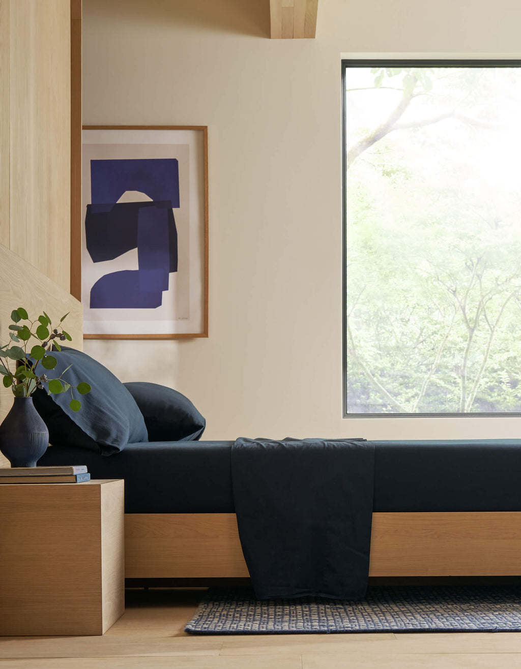 Minimalist bedroom featuring Cozy Earth’s Soft-Wash Cotton Sheet Set in dark blue, a wooden bed frame and nightstand, vase with greenery, abstract art on the wall, and a large window showcasing tree views and natural light.|Color:Eclipse