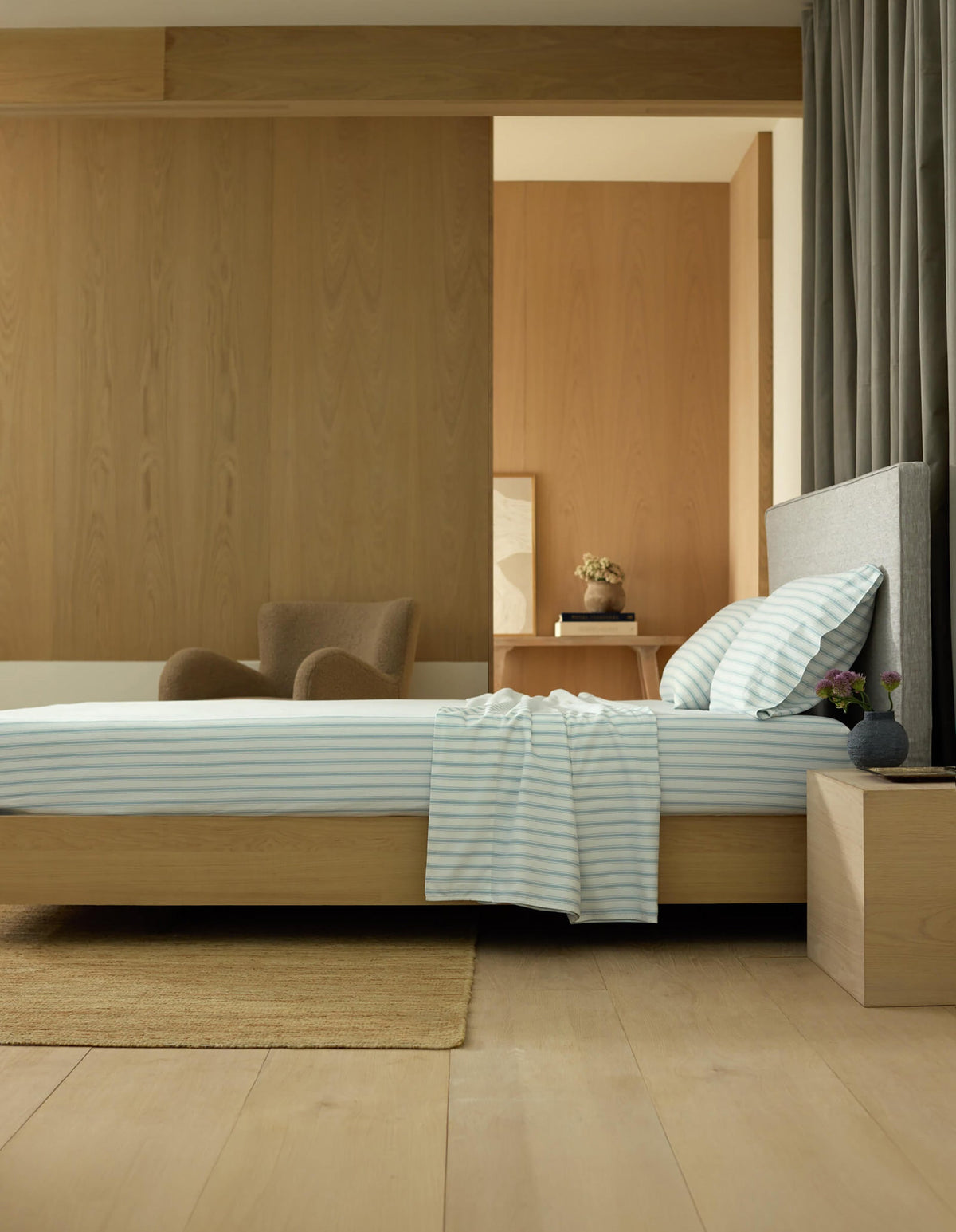 A modern, minimalist bedroom features a low wooden bed dressed in Cozy Earth's Soft-Wash Cotton Sheet Set, striped bedding, a wooden nightstand with a vase, an armchair, and wood-paneled walls and floor bathed in natural light.|Color:River and Sea Salt Stripe