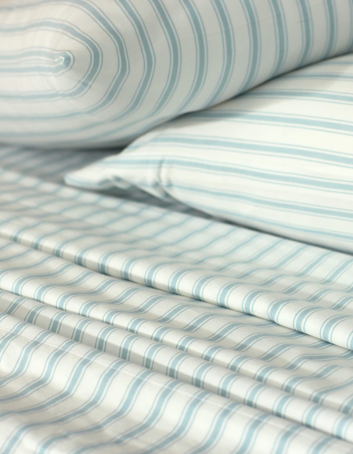Close-up of a neatly made bed showcasing the Cozy Earth Soft-Wash Cotton Sheet Set, including a pillow, fitted sheet, and flat sheet in a light blue and white striped pattern. The smooth fabric looks exceptionally soft.|Color:River and Sea Salt Stripe